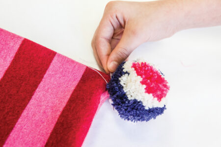 person sewing a blue red and white pom pom to the corner of red and pink fabric